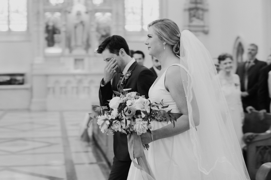 black and white photo of groom wiping his eyes as he walks up to the altar with his bride