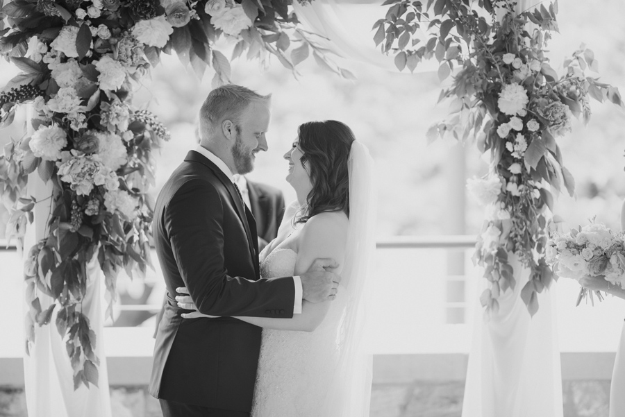 black and white of bride and groom holding each other and smiling under the floral arbor