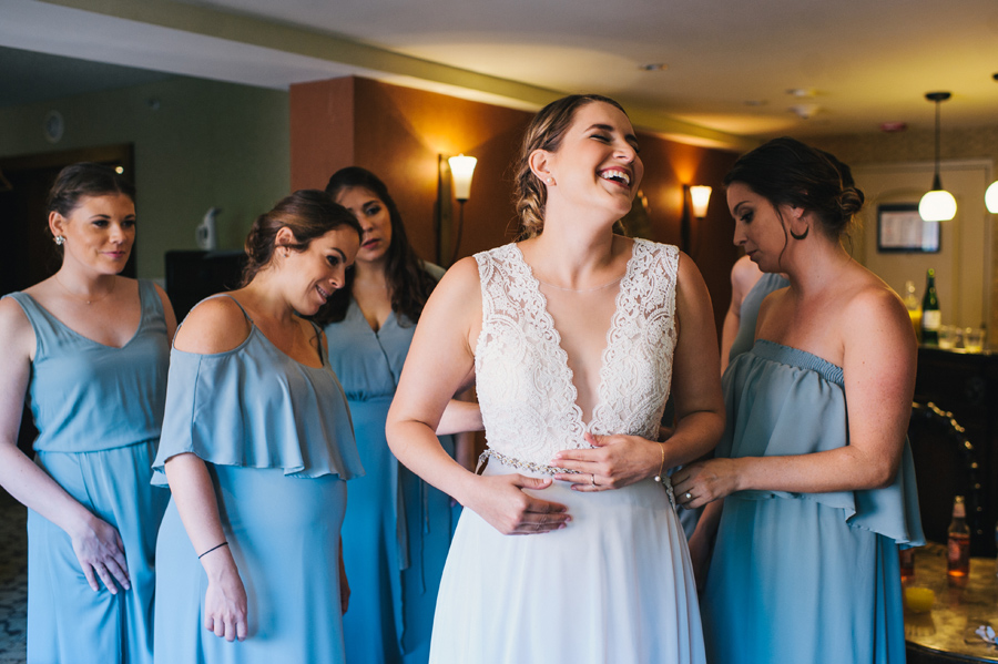 bride laughing while bridal party buttons the back of her dress