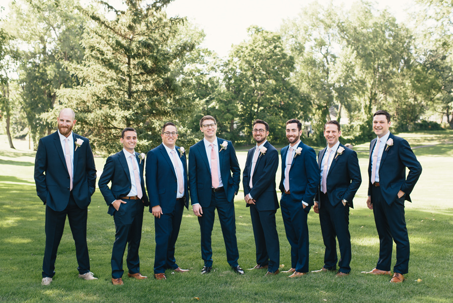 full length of groomsmen in navy suits with light pink ties standing in a park