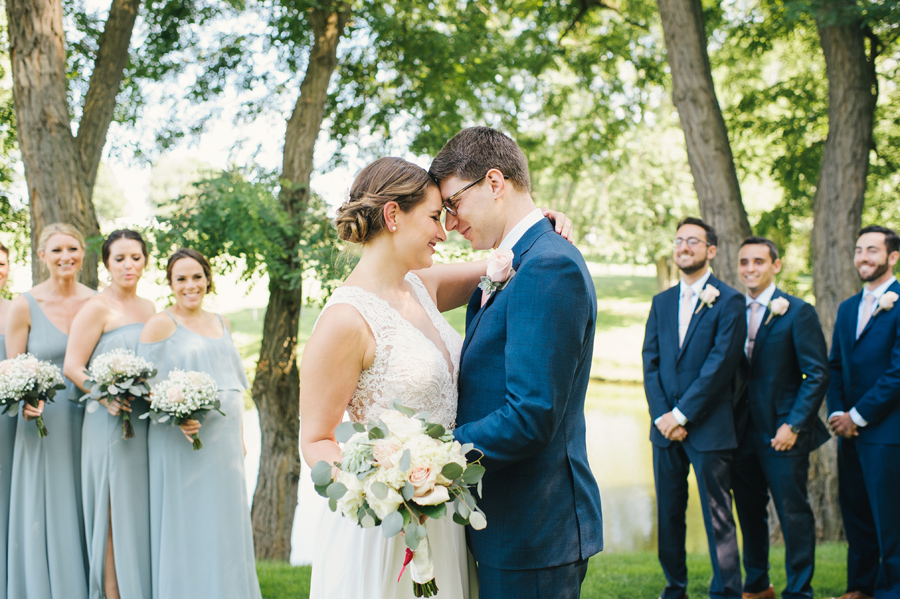 bride and groom smiling with foreheads together while wedding party looks on