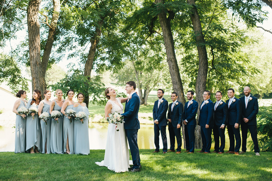 full length of bride laughing with the groom with wedding party lined up in the background and laughing