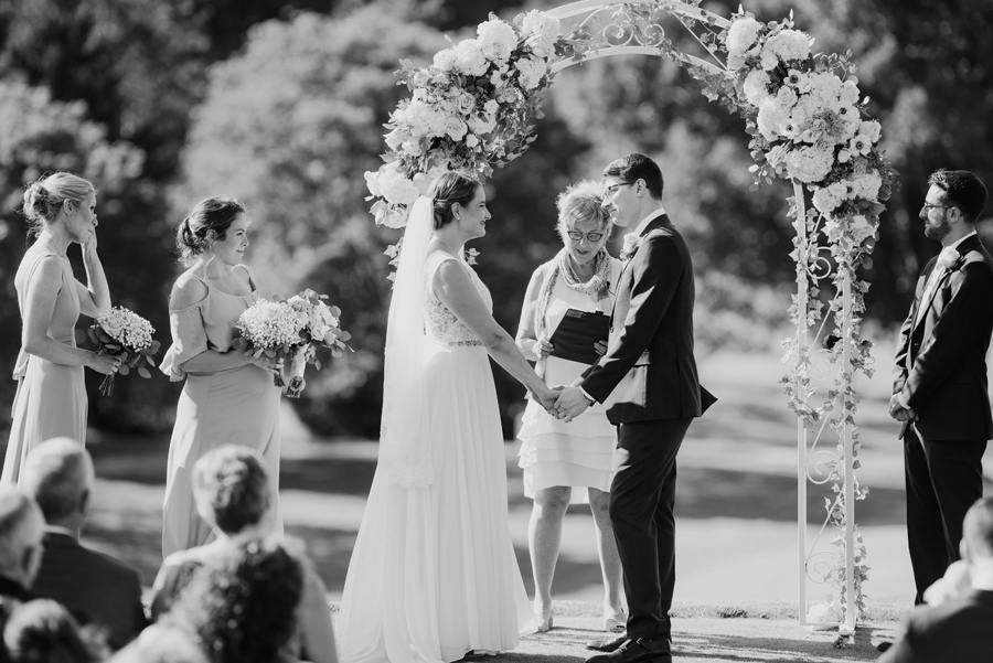 black and white of bride and groom holding hands under their floral arch during the wedding ceremony