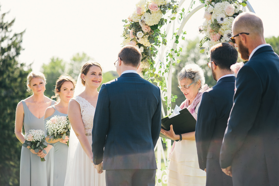 bride smiling at groom under a floral arch during their outdoor wedding ceremony