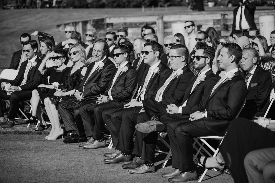 black and white shot of groomsmen and other seated guests watching the wedding ceremony on a golf course