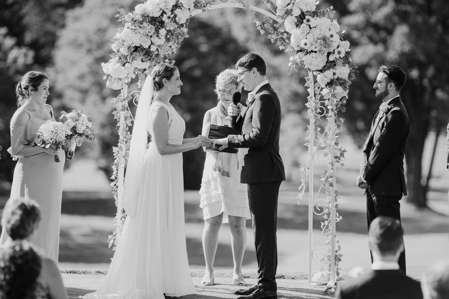 black and white shot of bride placing wedding band on groom's finger during their outdoor ceremony