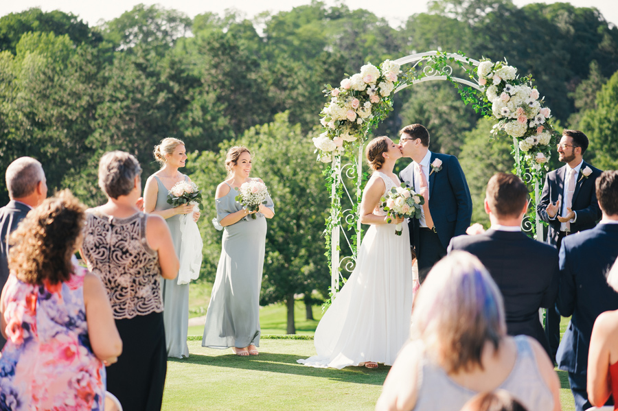 bride and groom kiss under their floral archway at the end of their outdoor country club wedding ceremony