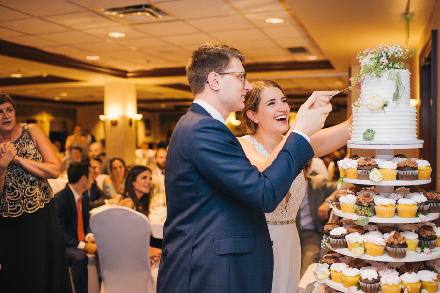 bride and groom cutting the two tier cake sitting atop a tower of cupcakes