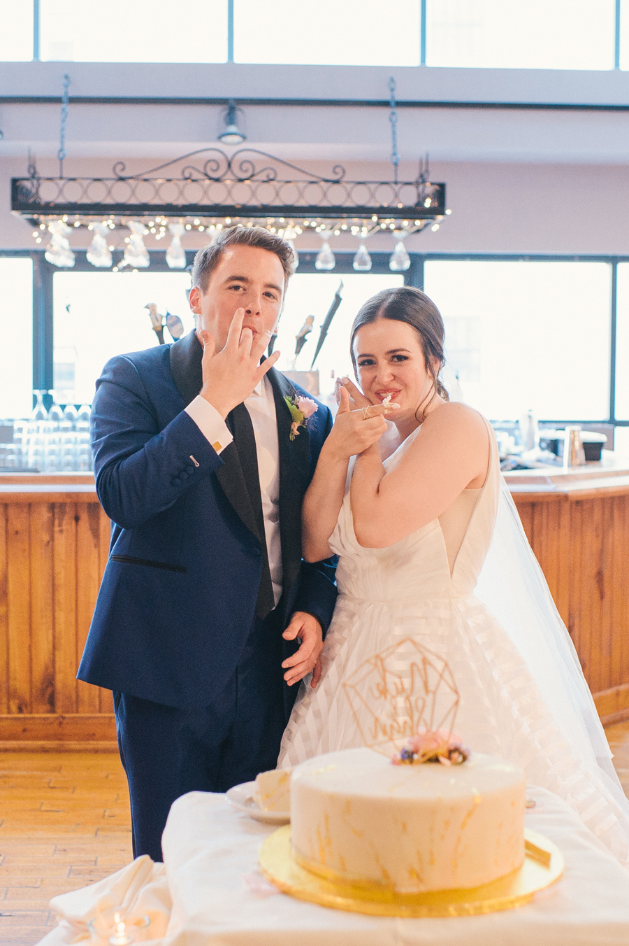 bride and groom looking at the camera and licking the cake frosting off their fingers