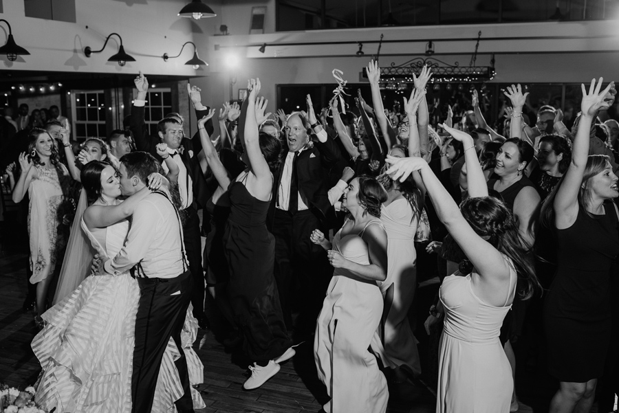 black and white of bride and groom kissing on the dance floor while wedding guest cheer around them