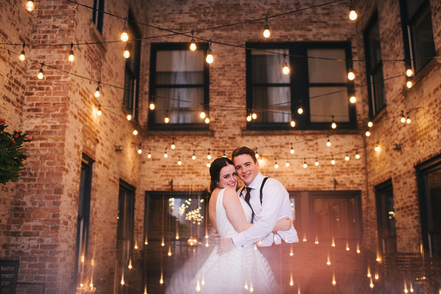 bride and groom holding each other close and smiling at the camera in the outdoor courtyard at night with many strands of twinkle lights