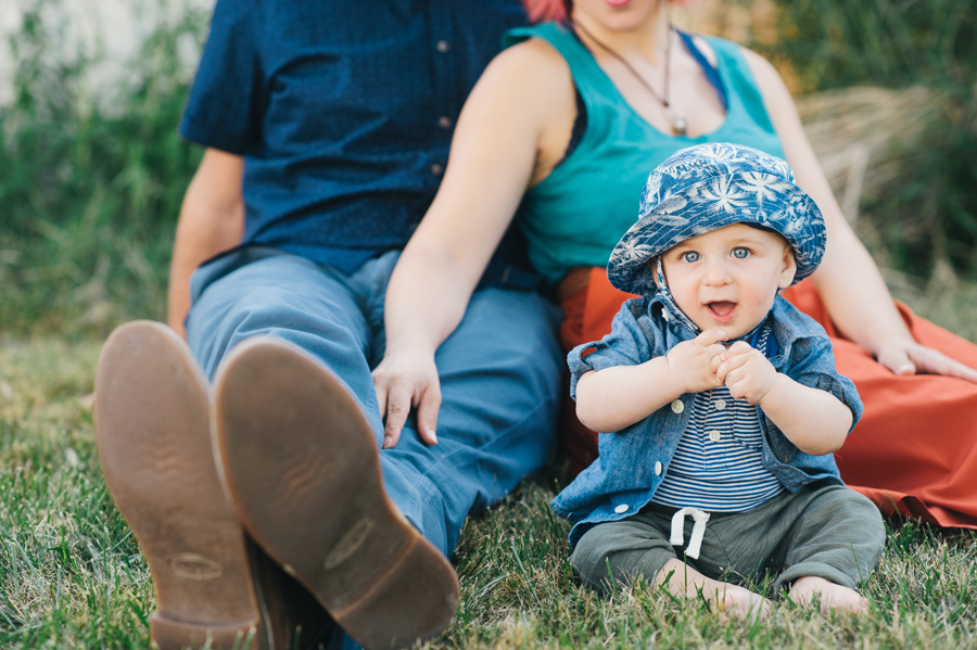 baby boy sitting in grass and looking coyly at camera with parents behind him