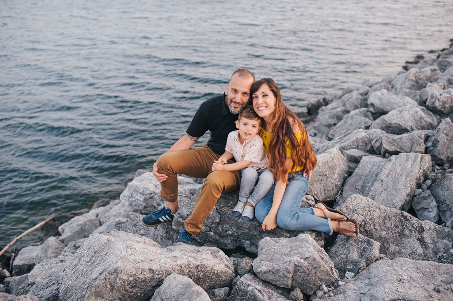 mom dad and son sitting on rocks at waters edge smiling at camera